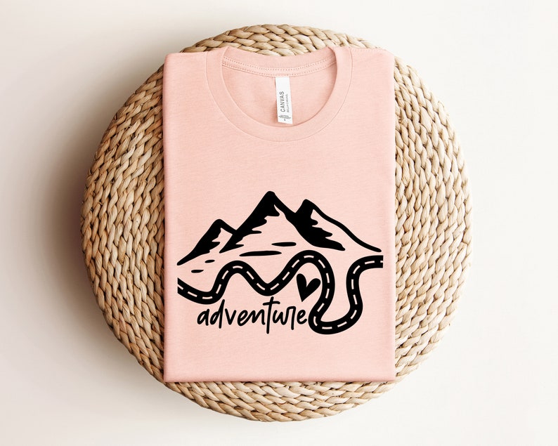 以下が含まれることがあります： 山と曲がりくねった道の黒いグラフィックが描かれたピンクのTシャツ。道の終わりにはハートマークがあります。グラフィックの下には「adventure」という文字があります。