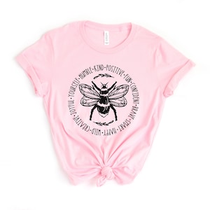 K&ouml;nnte beinhalten: Rosa T-Shirt mit einem schwarzen Bienen-Motiv und dem Text "WILD-CREATIVE-JOYFUL-YOURSELF-HUMBLE-KIND-POSITIVE-FUN-CONFIDENT-BRAVE-SMART-HAPPY" um die Biene herum.