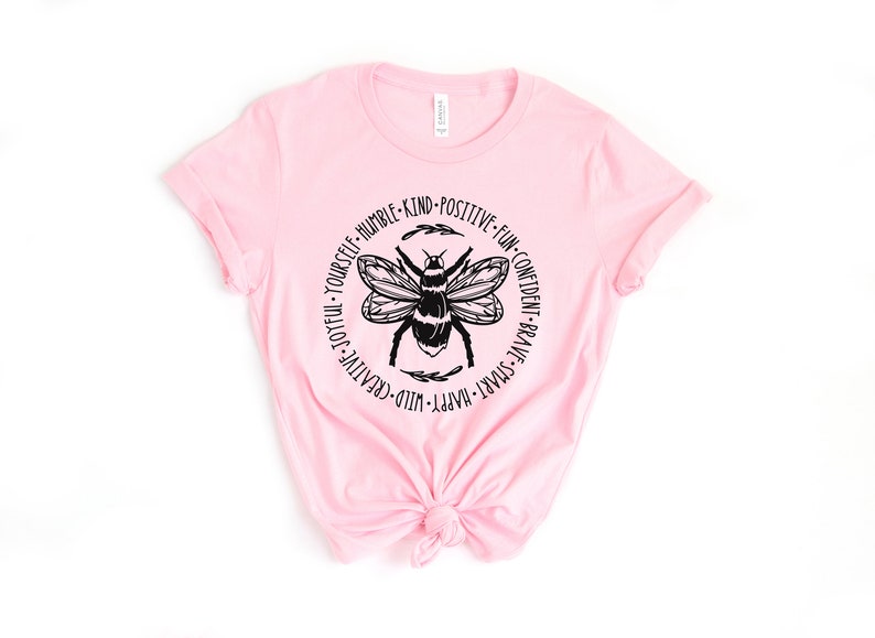 K&ouml;nnte beinhalten: Rosa T-Shirt mit einem schwarzen Bienen-Motiv und dem Text "WILD-CREATIVE-JOYFUL-YOURSELF-HUMBLE-KIND-POSITIVE-FUN-CONFIDENT-BRAVE-SMART-HAPPY" um die Biene herum.