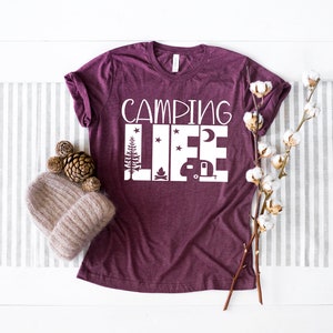 May include: A maroon t-shirt with the text "Camping Life" in white. The text is surrounded by images of a campfire, a camper, a moon, and stars.