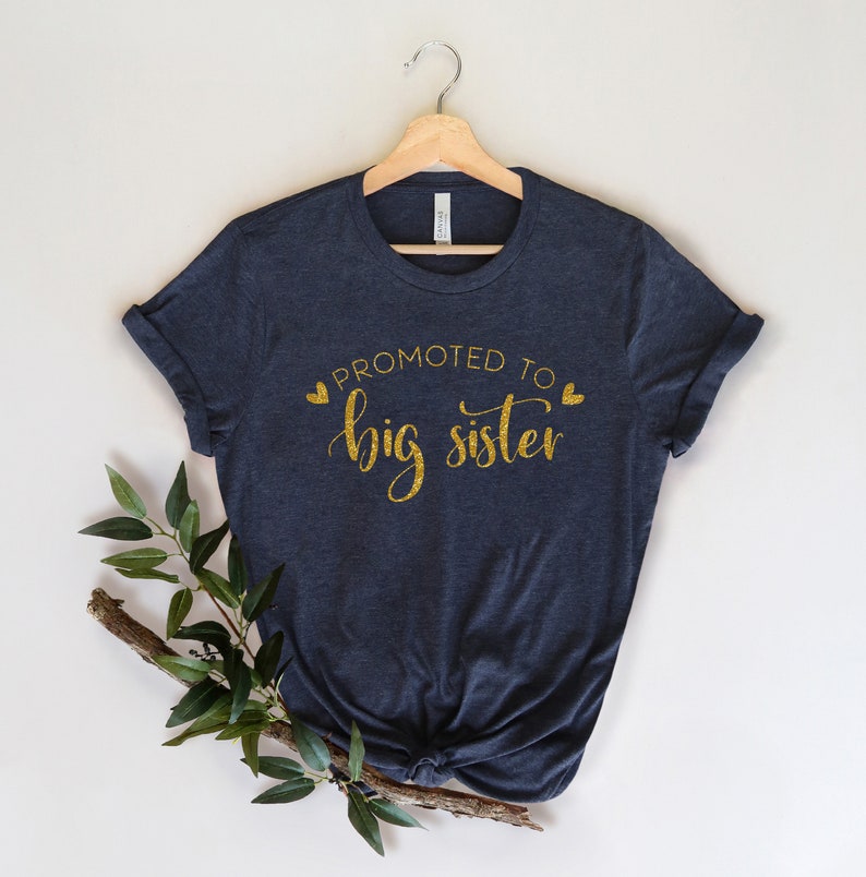 May include: A dark blue t-shirt with gold glitter text that reads "Promoted to Big Sister" with two small gold hearts on either side of the text.