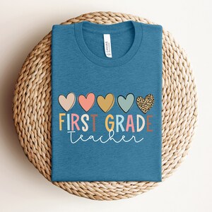 May include: A teal blue t-shirt with the text "FIRST GRADE teacher" in colorful letters and a row of five hearts above the text. The hearts are pink, red, yellow, blue, and leopard print.
