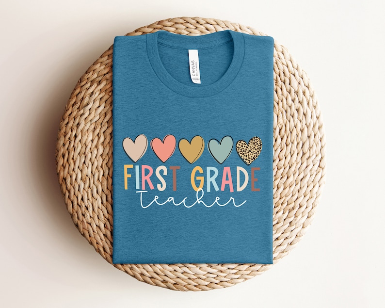 May include: A teal blue t-shirt with the text "FIRST GRADE teacher" in colorful letters and a row of five hearts above the text. The hearts are pink, red, yellow, blue, and leopard print.