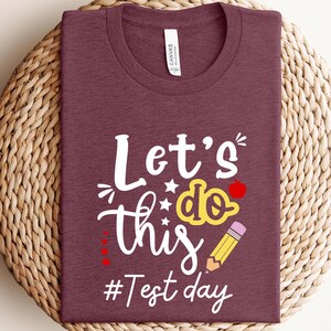 May include: Burgundy t-shirt with white text that reads "Let's do this #Test day". The text is decorated with a yellow star, a yellow pencil, and a red apple.