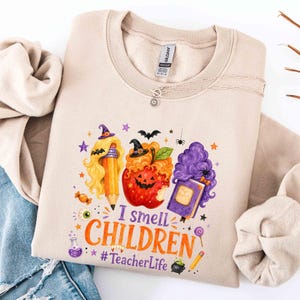 May include: A sand-colored crewneck sweatshirt featuring a graphic print with a pencil, a jack-o'-lantern, and a book, all wearing witch hats. The text reads I smell CHILDREN and #TeacherLife, surrounded by small stars, bats, and a potion bottle.