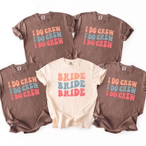 Puede incluir: Cinco camisetas con mangas enrolladas. Cuatro son marrones con el texto "I DO CREW" en naranja, azul y rosa. Una es de color crema con "BRIDE" en los mismos colores. Ropa para la fiesta de bodas.
