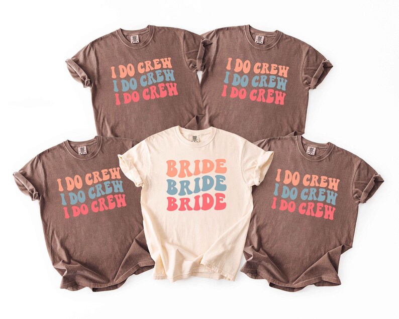 Puede incluir: Cinco camisetas con mangas enrolladas. Cuatro son marrones con el texto "I DO CREW" en naranja, azul y rosa. Una es de color crema con "BRIDE" en los mismos colores. Ropa para la fiesta de bodas.