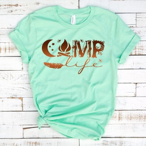 May include: A mint green t-shirt with a copper-colored graphic that says "Camp Life". The graphic includes a crescent moon, a campfire, and a feather.