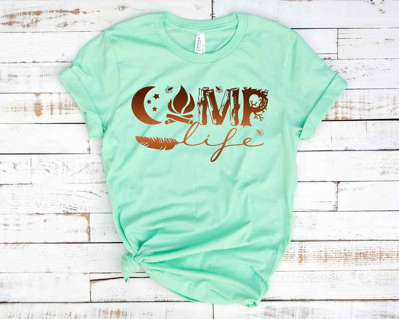 May include: A mint green t-shirt with a copper-colored graphic that says "Camp Life". The graphic includes a crescent moon, a campfire, and a feather.
