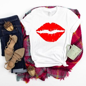 May include: White t-shirt with a red lipstick print. The shirt is laid out on a plaid blanket with a pair of brown sandals and blue jeans.
