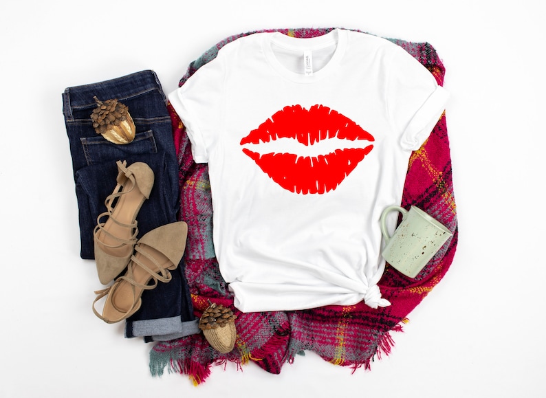 May include: White t-shirt with a red lipstick print. The shirt is laid out on a plaid blanket with a pair of brown sandals and blue jeans.