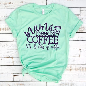 Puede incluir: Una camiseta verde claro con el texto "mama needs coffee lots & lots of coffee" en letras de color morado oscuro. Un icono de taza de caf&eacute; est&aacute; al lado de la palabra "coffee".