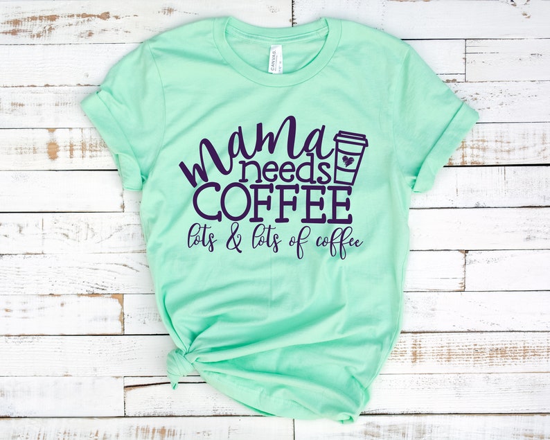 Puede incluir: Una camiseta verde claro con el texto "mama needs coffee lots & lots of coffee" en letras de color morado oscuro. Un icono de taza de caf&eacute; est&aacute; al lado de la palabra "coffee".