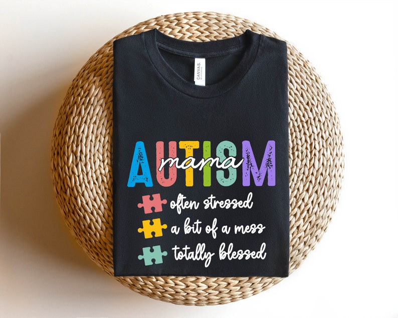K&ouml;nnte beinhalten: Schwarzes T-Shirt mit dem Text "AUTISM mama" in Regenbogenfarben. Unter dem Text befinden sich drei Puzzle-Teile mit dem Text "oft gestresst", "ein bisschen ein Chaos" und "total gesegnet".