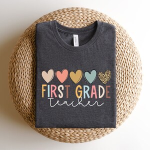 May include: A dark grey t-shirt with the text "FIRST GRADE teacher" in colorful letters and a row of five hearts above the text. The hearts are pink, orange, yellow, blue, and leopard print.