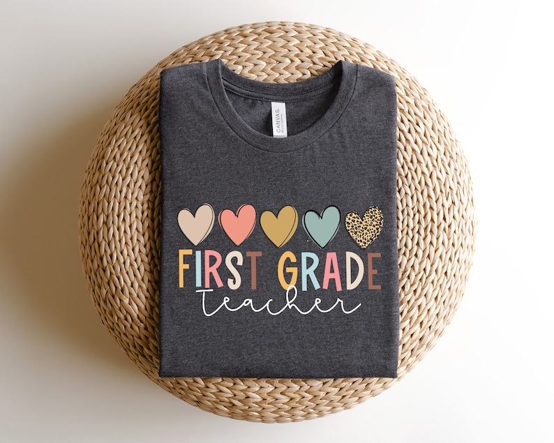 May include: A dark grey t-shirt with the text "FIRST GRADE teacher" in colorful letters and a row of five hearts above the text. The hearts are pink, orange, yellow, blue, and leopard print.