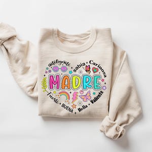 Puede incluir: Una sudadera beige con un dise&ntilde;o gr&aacute;fico colorido que dice "MADRE" rodeado de palabras en espa&ntilde;ol, incluyendo "inteligente", "sabia", "cari&ntilde;osa", "fuerte", "buena", "bella" y "valiente".