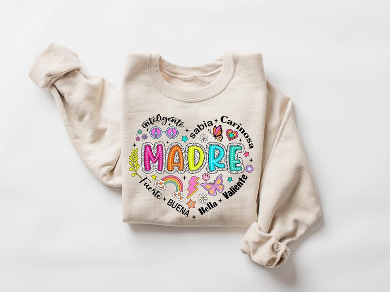 Puede incluir: Una sudadera beige con un dise&ntilde;o gr&aacute;fico colorido que dice "MADRE" rodeado de palabras en espa&ntilde;ol, incluyendo "inteligente", "sabia", "cari&ntilde;osa", "fuerte", "buena", "bella" y "valiente".
