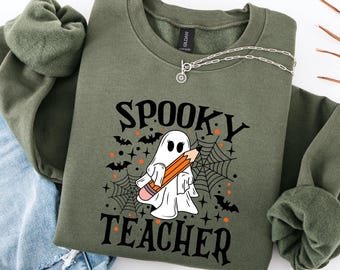 May include: Olive green sweatshirt with the words "Spooky Teacher" in black, featuring a white ghost holding an orange pencil. Black bats, spiderwebs, and orange stars decorate the design. A silver necklace is draped across the sweatshirt.