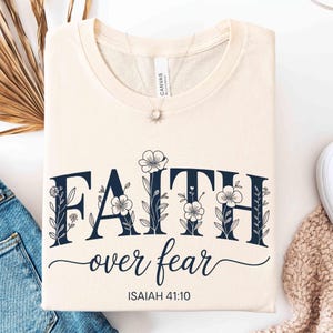 May include: Cream-colored t-shirt with the words "FAITH over fear" in navy blue, adorned with floral accents. The text "ISAIAH 41:10" is below. The shirt is folded neatly, showcasing the design.