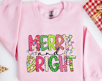 Puede incluir: Una sudadera rosa claro con las palabras "MERRY and BRIGHT" en letras coloridas y estampadas. El diseño incluye luces navideñas y patrones festivos, perfecto para la temporada navideña.