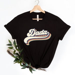 Może przedstawiać: Czarny T-shirt z grafiką w stylu retro, na kt&oacute;rej widnieje napis "Dada" w kolorach tęczy.