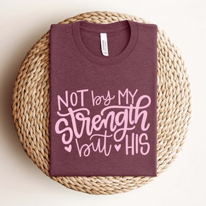 Op de afbeelding: Een maroon t-shirt met de tekst "NOT by MY strength but HIS" in roze letters. Het shirt is gevouwen en geplaatst op een geweven stro mat.