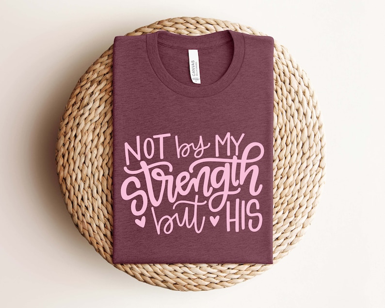Op de afbeelding: Een maroon t-shirt met de tekst "NOT by MY strength but HIS" in roze letters. Het shirt is gevouwen en geplaatst op een geweven stro mat.