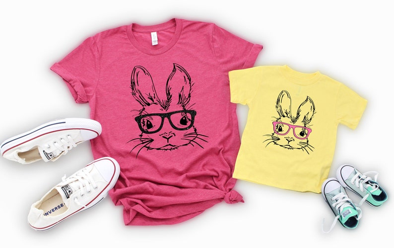 May include: Two t-shirts featuring a black and white illustration of a bunny wearing pink glasses. The larger shirt is pink and the smaller shirt is yellow. Both shirts are laid out on a white background with white sneakers.