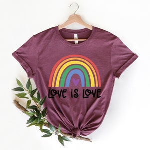 Puede incluir: Una camiseta de color granate con un arco&iacute;ris y el texto "LOVE IS LOVE" impreso en la parte delantera.