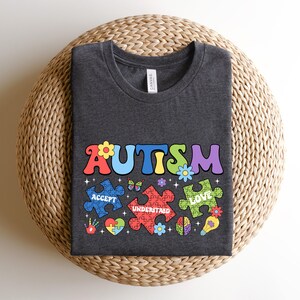 May include: A dark gray t-shirt with the word "AUTISM" in colorful letters. The letters are surrounded by puzzle pieces with the words "ACCEPT", "UNDERSTAND", and "LOVE" on them.