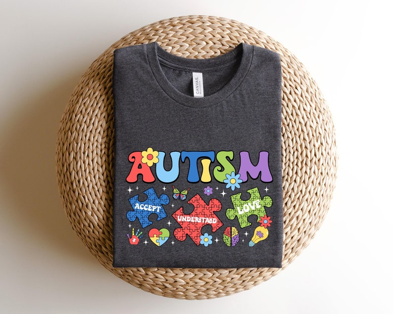 May include: A dark gray t-shirt with the word "AUTISM" in colorful letters. The letters are surrounded by puzzle pieces with the words "ACCEPT", "UNDERSTAND", and "LOVE" on them.
