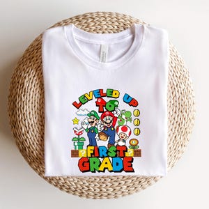 Puede incluir: Camiseta blanca con un gr&aacute;fico colorido de Mario, Luigi y otros personajes de la serie de videojuegos Super Mario. El texto "LEVELED UP TO FIRST GRADE" se muestra en una fuente vibrante y multicolor.