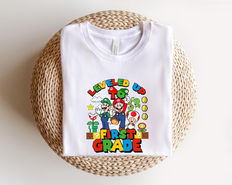 Puede incluir: Camiseta blanca con un gr&aacute;fico colorido de Mario, Luigi y otros personajes de la serie de videojuegos Super Mario. El texto "LEVELED UP TO FIRST GRADE" se muestra en una fuente vibrante y multicolor.