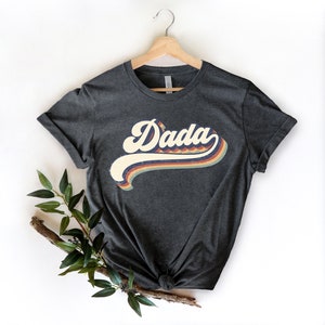 Może przedstawiać: Szary t-shirt z grafiką w stylu retro, na kt&oacute;rej widnieje napis "Dada" w kolorowym tęczowym wzorze.