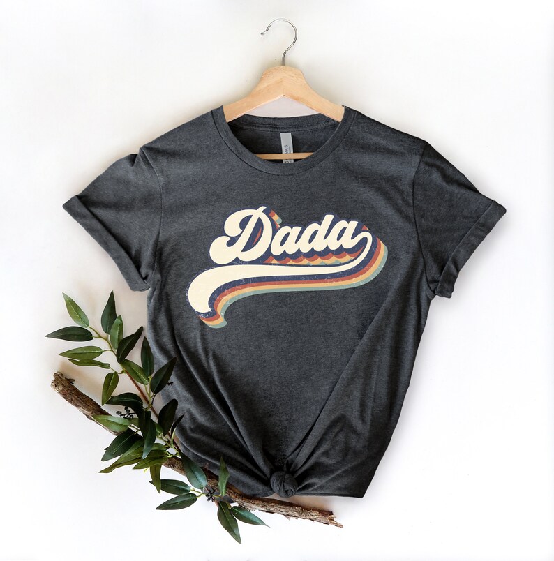 Może przedstawiać: Szary t-shirt z grafiką w stylu retro, na kt&oacute;rej widnieje napis "Dada" w kolorowym tęczowym wzorze.
