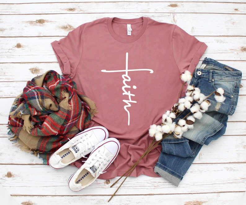 May include: A dusty rose colored t-shirt with a white cross and the word "faith" written vertically on the cross. The t-shirt is laying on a white wooden surface with a plaid scarf, a pair of white sneakers, a pair of blue jeans, and a sprig of cotton.
