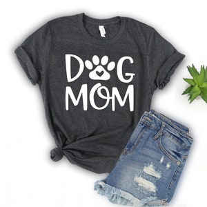 Pode incluir: Uma camiseta cinza escura com o texto "DOG MOM" em letras brancas. Uma pegada de pata branca com um pequeno cora&ccedil;&atilde;o dentro est&aacute; entre as palavras "DOG" e "MOM".
