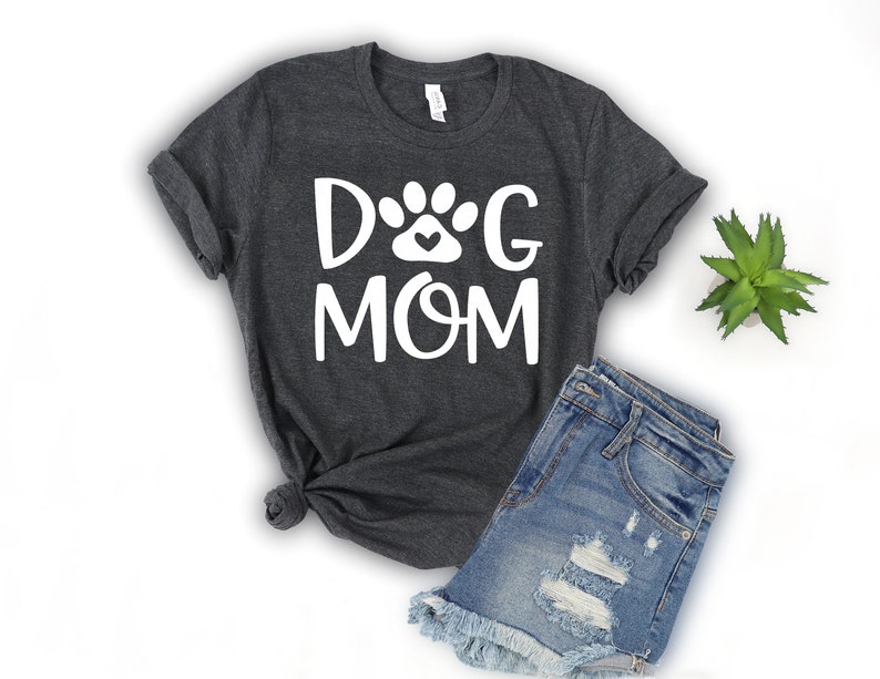 Pode incluir: Uma camiseta cinza escura com o texto "DOG MOM" em letras brancas. Uma pegada de pata branca com um pequeno cora&ccedil;&atilde;o dentro est&aacute; entre as palavras "DOG" e "MOM".