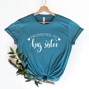 May include: Teal blue t-shirt with white text that reads "Promoted to big sister" with two small hearts on either side of the text.