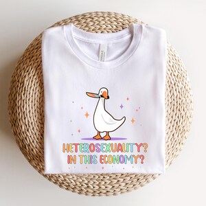 K&ouml;nnte beinhalten: Wei&szlig;es T-Shirt mit einer Cartoon-Gans, die ein regenbogenfarbenes Klebeband &uuml;ber ihrem Schnabel tr&auml;gt. Der Text "Heterosexuality? In this economy?" ist in Regenbogenfarben unter der Gans gedruckt.