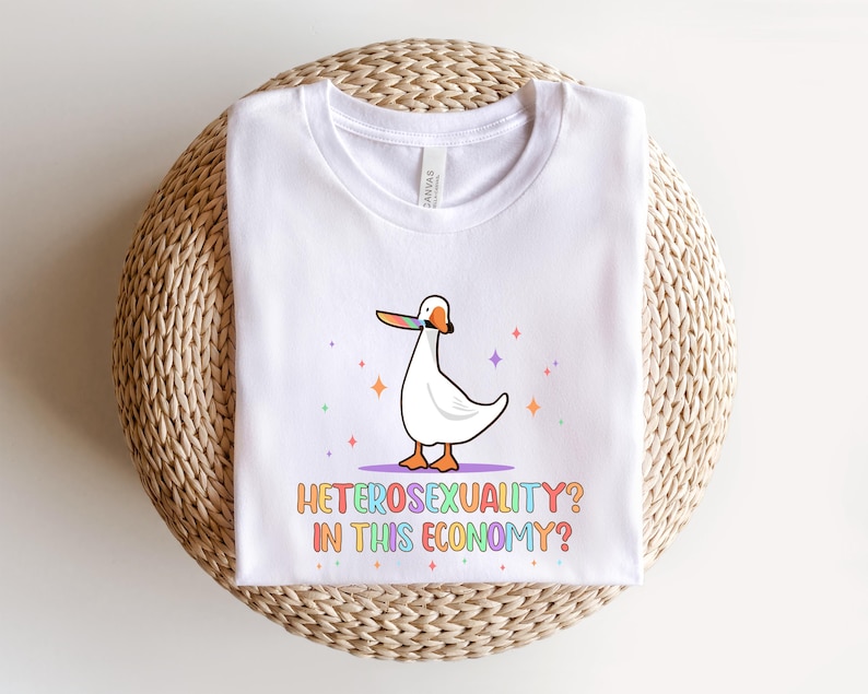 K&ouml;nnte beinhalten: Wei&szlig;es T-Shirt mit einer Cartoon-Gans, die ein regenbogenfarbenes Klebeband &uuml;ber ihrem Schnabel tr&auml;gt. Der Text "Heterosexuality? In this economy?" ist in Regenbogenfarben unter der Gans gedruckt.