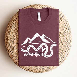 以下が含まれることがあります： 山と曲がりくねった道の白いグラフィックが描かれた、マルーンのTシャツ。Tシャツには「冒険」という言葉が書かれています。