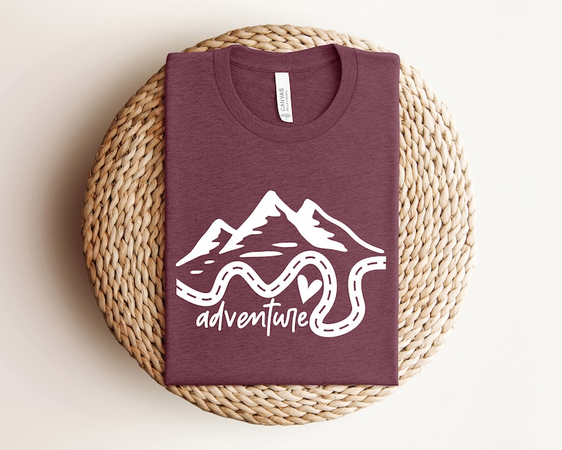 以下が含まれることがあります： 山と曲がりくねった道の白いグラフィックが描かれた、マルーンのTシャツ。Tシャツには「冒険」という言葉が書かれています。