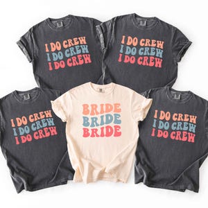 Puede incluir: Cinco camisetas, cuatro en gris oscuro y una en crema. Las camisetas grises oscuras tienen el texto "I DO CREW" en naranja, azul y rojo. La camiseta crema dice "BRIDE" en los mismos colores. Todas las camisetas son de manga corta.