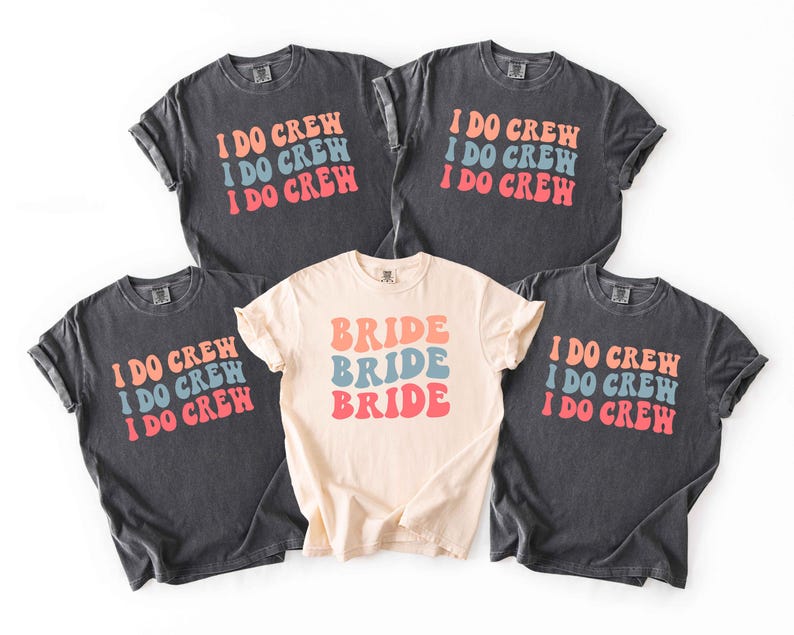 Puede incluir: Cinco camisetas, cuatro en gris oscuro y una en crema. Las camisetas grises oscuras tienen el texto "I DO CREW" en naranja, azul y rojo. La camiseta crema dice "BRIDE" en los mismos colores. Todas las camisetas son de manga corta.
