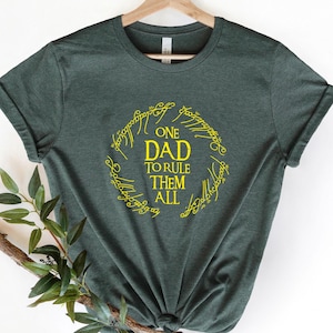 May include: Dark green t-shirt with a yellow graphic. The graphic features the text "ONE DAD TO RULE THEM ALL" inside a circular design with stylized lettering. The shirt is displayed on a wooden hanger.