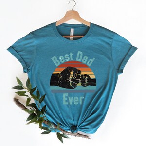 May include: Teal blue t-shirt featuring a vintage-style graphic of two fists bumping. The graphic includes a sunset with red, orange, yellow, and blue stripes. The text "Best Dad Ever" is printed below the graphic.