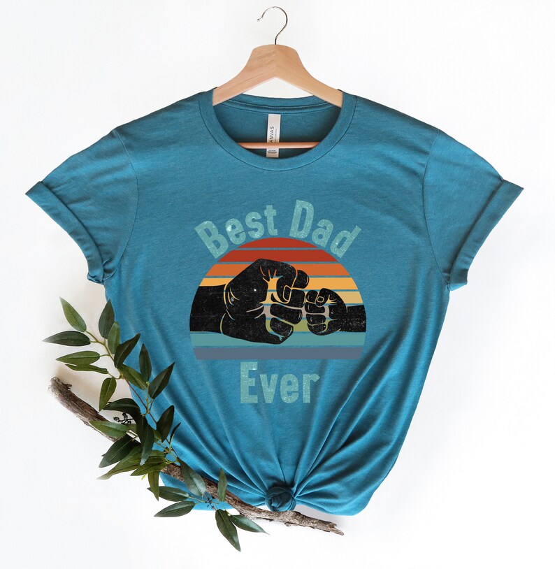 May include: Teal blue t-shirt featuring a vintage-style graphic of two fists bumping. The graphic includes a sunset with red, orange, yellow, and blue stripes. The text "Best Dad Ever" is printed below the graphic.
