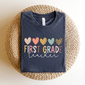 May include: A dark heather grey t-shirt with the text "FIRST GRADE teacher" in colorful letters and a row of five hearts in various colors and patterns above the text.